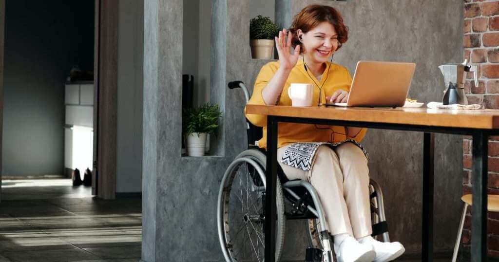 A woman in a wheelchair is waving to her laptop during a virtual coffee chat. She has a cup of coffee next to her.